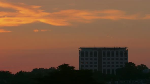 Zooming Out timelapse of a tall building and a silhouette of trees with the sun arching into shot ov