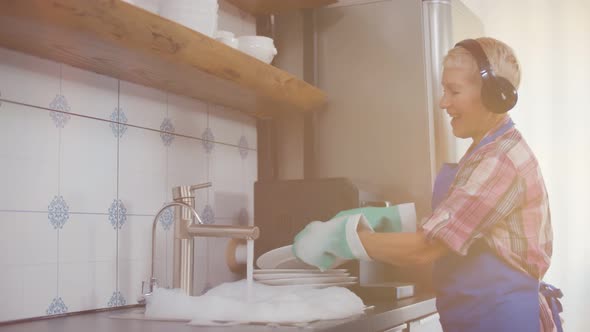 Positive Senior Woman Cleaning Dirty Plates and Listening To Music in Headset