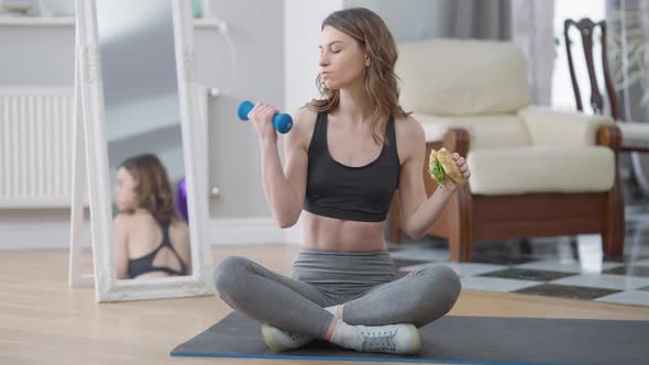 Front View of Slim Woman Eating Unhealthy Food Lifting Dumbbell Sitting on Exercise Mat