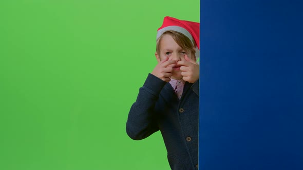 Child Boy in a Christmas Hat Comes From Behind the Wall and Shows Tongue on a Green Screen