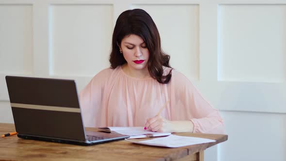 Pretty Young Lady Sitting at Desk and Typing on Laptop and Writing Tasks for the Day in a Notebook