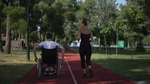 Live Camera Follows Man Rolling Wheelchair As Woman Jogging on Sports Track Outdoors