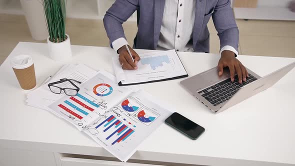 Office Manager Sitting at Working Table and Writing Down Important Marks from Laptop Into the Report