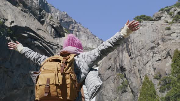 Smiling Woman with Tourist Backpack Raises Arms Into Air Taking Off Pink Hat