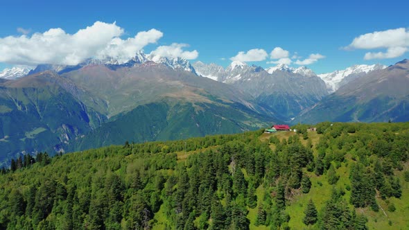 Aerial View of a Small House in the Forest Against the Background of Snowcapped Mountain Peaks