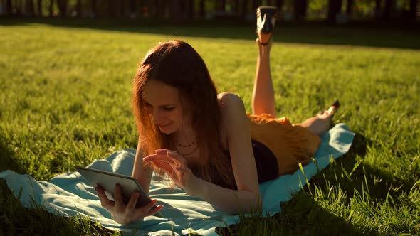 Female Sitting in the Park Among the Trees and Use the Internet on Tablet.