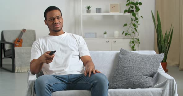 Afro American Young Sad Guy Black Ethnic Man in White Tshirt Sits on Gray Sofa in Cozy Living Room