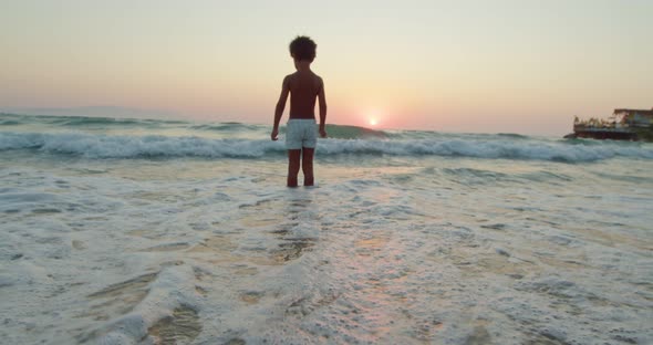 AfricanAmerican Boy Stands in the Waves of the Sea and Looks at the Departing Sun a View From the