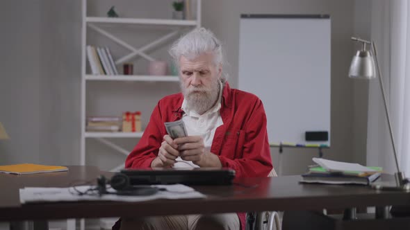 Bearded Senior Handicapped Freelancer Counting Money Looking at Camera Thinking