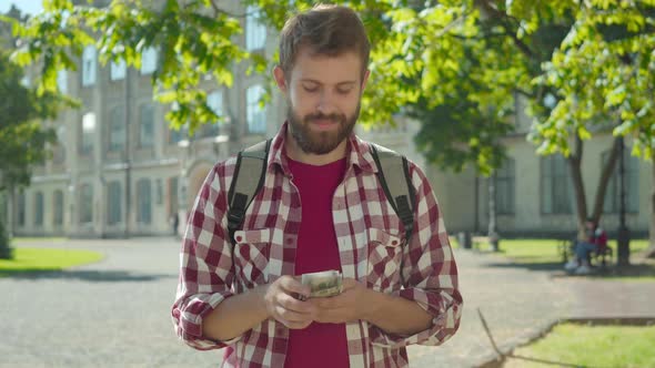 Happy Satisfied Caucasian Man Counting Money on University Yard and Looking Away. Portrait of Young