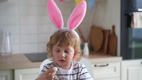 Cute two-year-old boy in bunny ears paint Easter eggs with multi-color paints sitting at the table