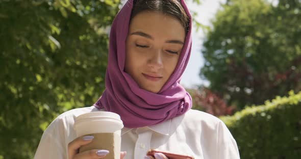 Beautiful Young Woman Drink Coffee and Looking at Smartphone, Wearing Traditional Headscarf