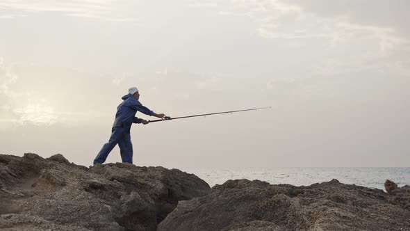 Old fisherman standing on sea side rocks and fishing against the sunset