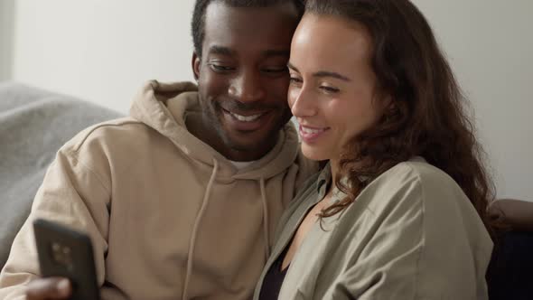 Relaxed Young Couple At Home Sitting On Sofa Checking Social Media On Mobile Phone