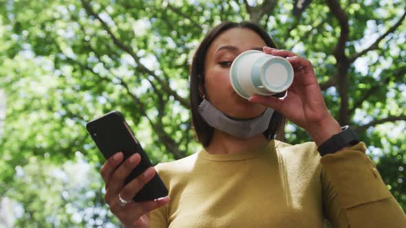 African american woman wearing face mask using smartphone drinking coffee in park