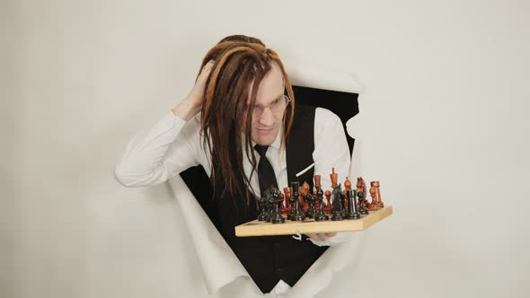 Young Man in Glasses with Dreadlocks Plays Chess in Hole of White Background