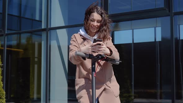 A Young Student Girl in a Sweater Jeans and a Coat Stands with an Electric Scooter Near a Stylish