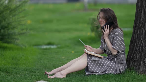 a Brunette in a Striped Dress Glasses and Barefoot is Sitting Under a Tree with a Book on Knees