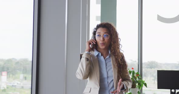 Biracial businesswoman sitting by desk and talking on smartphone in modern interiors