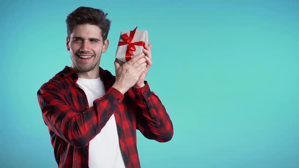 Handsome man holding gift box on blue studio background smiles to camera. 