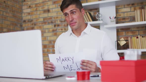 Caucasian man making video call using laptop holding handwritten sign making marriage proposal