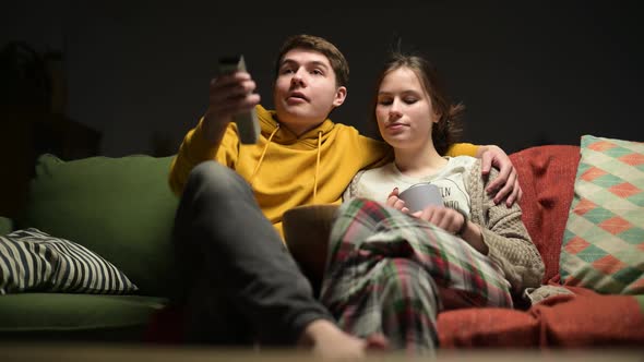 A young couple sits down on the sofa in the evening and turns on the TV