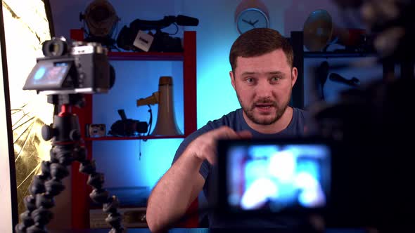 Man Blogger Sits at Table with Two Cameras  Red Monitor and Conducts Video Blog