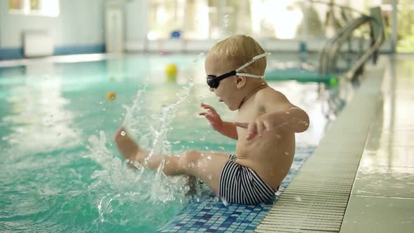 A Little Boy with Blond Hair Sits By the Pool with His Feet in the Water