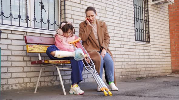 Mom Talking on Phone Daughter with Crutches on Bench in Yard