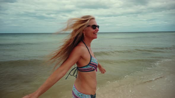 Laughing, playful mature woman in bikini and sunglasses turns around and runs on the beach.