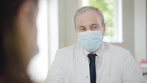 Mid-adult Doctor in Face Mask Consulting Patient on Covid-19 Pandemic. Blurred Patient at Front