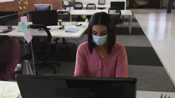 Mixed race businesswoman wearing face mask sitting at desk using computer