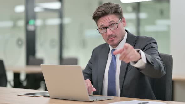 Middle Aged Businessman with Laptop Pointing at the Camera 