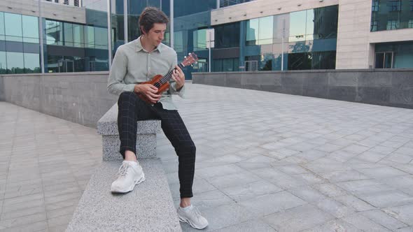Young Attractive Guy in Casual Clothes and White Sneakers Sitting Outdoors Playing on Ukulele