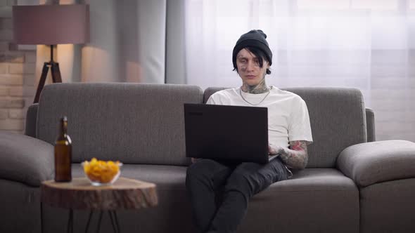 Confident Concentrated Tattooed Man Sitting on Couch Surfing Internet on Laptop