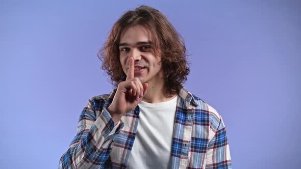 Smiling Man with Long Hairstyle Holding Finger on Lips Over Violet Background