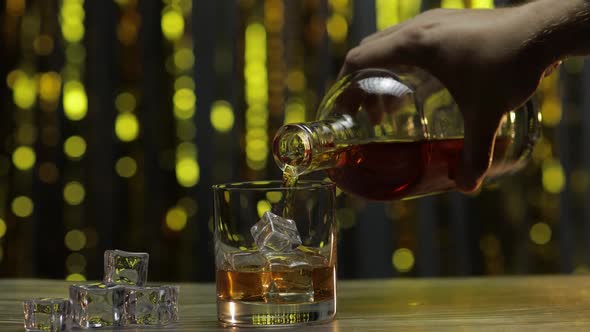 Pouring of Golden Whiskey, Cognac or Brandy From Bottle Into Glass with Ice Cubes. Shiny Background