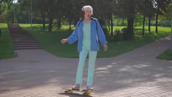 Wide Shot Portrait of Happy Confident Female Retiree Standing with Skateboard in Sunlight Gesturing