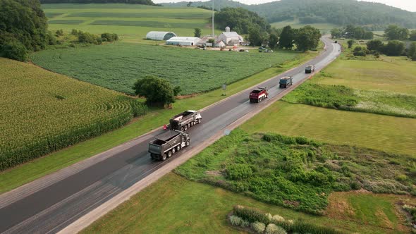 Road construction crew working with dump trucks on a rural highway with farm fields on each side.