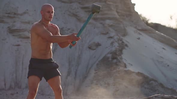 Bald Man Strongman Hits a Hammer on a Huge Wheel in the Sandy Mountains in Slow Motion. Strength and