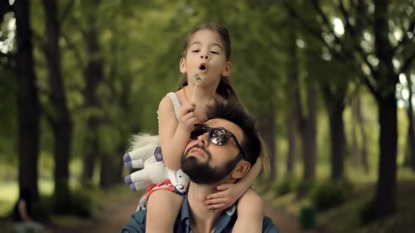 Father's Day.  Baby Girl And Father Blowing Dandelion. Child Daughter Sitting On Daddy Neck.