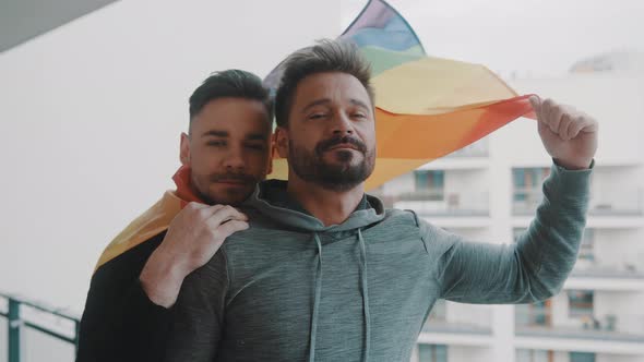 Young Excited Gay Male Couple Hugging on the Balcony Covered with Rainbow Flag