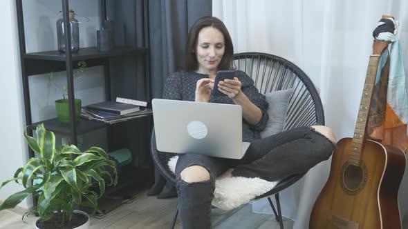 Attractive young woman sitting on cozy chair in jeans and shirt using laptop computer for work