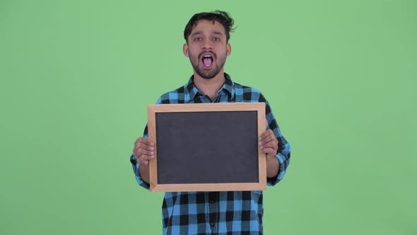 Happy Young Bearded Persian Hipster Man Holding Blackboard and Looking Surprised