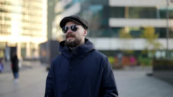 a Mustachioed and Bearded Man in a Cap and Dark Glasses Walks Against the Background of a Blurred