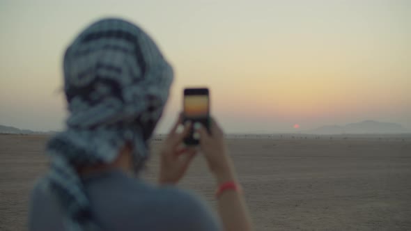 Woman in Checkered Keffiyeh Taking Picture of Sunrise on Mobile Phone Standing in the Desert