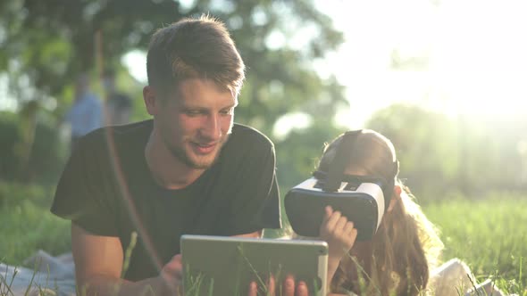 Caucasian Young Man Using Tablet Lying with Little Girl Wearing VR Headset in Sunbeam