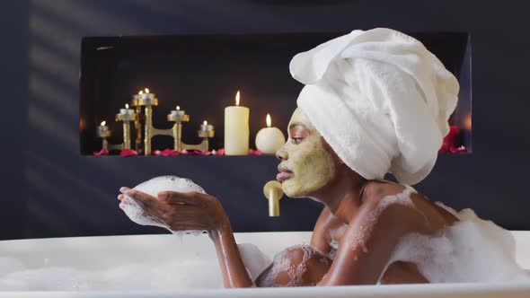 African american woman with towel and mask taking bath and blowing foam in bathroom