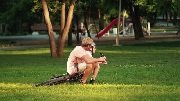 A happy father is sitting with his son on the grass in the park after riding a bicycle at summer