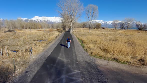 Mature, balding man walking with a backpack down a nature trail with the snow-capped mountains in th